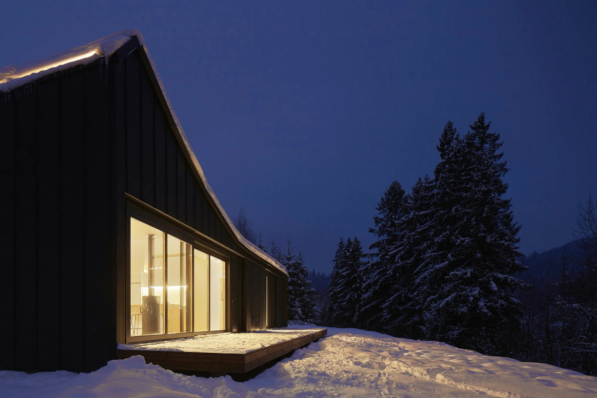 Moderne dunkle Hütte zwischen verschneiten Fichten zur blauen Stunde, warmes Licht leuchtet aus den großen Fenstern